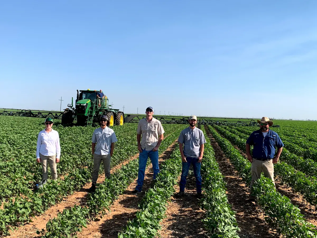 5 men and woman stand in a line among crop rows and farm equipment